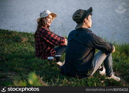 Cheerful Young backpacker couple sitting on grass and looking forward over lake in early morning and making fresh coffee grinder while c&ing trip on summer vacation