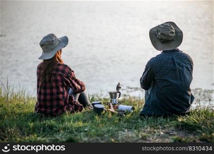 Cheerful Young backpacker couple sitting on grass and looking forward over lake in early morning and making fresh coffee grinder while c&ing trip on summer vacation