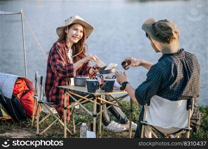 Cheerful Young backpacker couple wearing trekking hat sitting near lake with coffee and breakfast and making fresh coffee grinder while c&ing trip on summer vacation