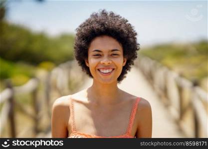 Cheerful young black woman with curly hair smiling and looking at camera while standing on wooden path on sunny summer day. Happy African American woman on boardwalk