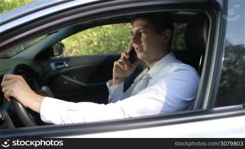 Cheerful young business executive in white shirt and tie talking on mobile phone as he sits in his car and holding steering wheel with one hand. Successful businessman communicating with business partner on smartphone while driving car. Slo mo. Stabilized shot.