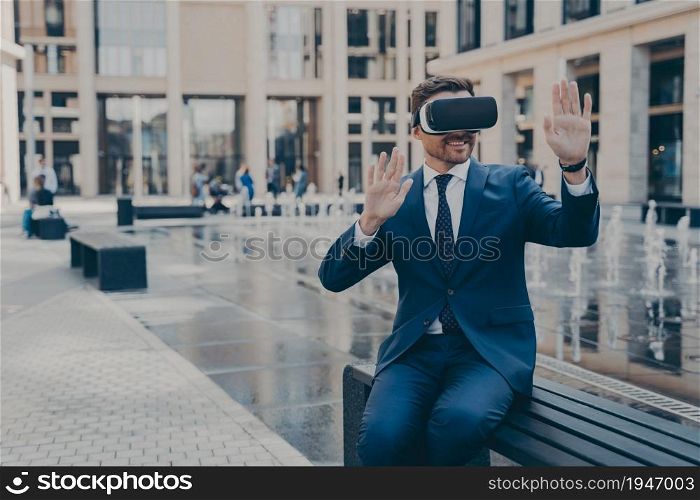 Cheerful young business owner in formal suit reviewing projects in virtual reality, using portable VR device goggles while sitting on bench, gesturing with hands and smiling, testing simulation app. Young business owner in blue formal suit reviewing projects in virtual reality