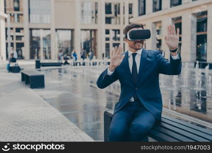 Cheerful young business owner in formal suit reviewing projects in virtual reality, using portable VR device goggles while sitting on bench, gesturing with hands and smiling, testing simulation app. Young business owner in blue formal suit reviewing projects in virtual reality