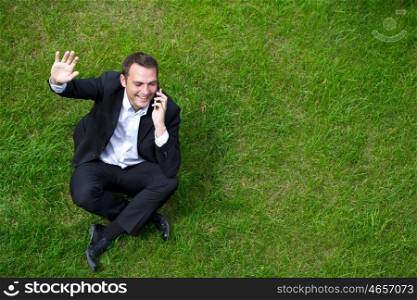Cheerful young businessman calling by phone sitting on green grass