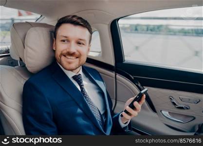 Cheerful young corporate executive in elegant expensive tuxedo rides in luxury car with company driver to financial meeting while checking market stock prices and indices on his cellphone. Corporate executive in elegant expensive tuxedo rides in luxury car