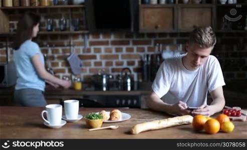 Cheerful young couple in love having breakfast together in the kitchen. Handsome hipster sitting at table and surfing the net with smart phone. Beautiful brunette girl bringing mason jars with orange juice, gently embracing and kissing her boyfriend.