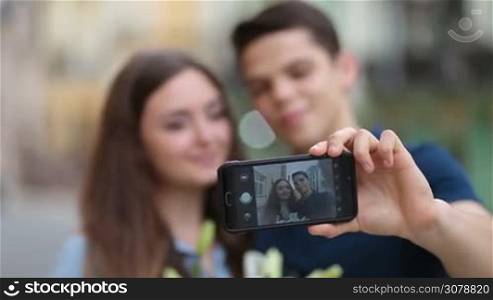 Cheerful young couple in love taking selfie on mobile phone during their travel on vacation. Focus on smartphone with blurry people on background. Closeup.