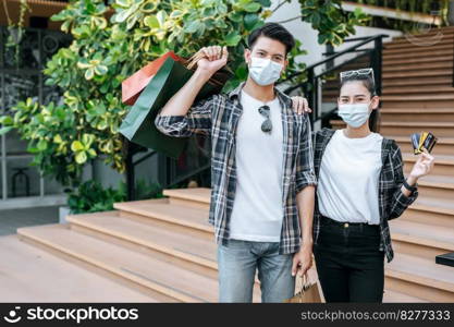 Cheerful Young couple in mask holding multiple paper shopping bag going down stairs of large shopping mall and pretty woman show credit card in hand,  Lifestyle and shopping concept, copy space