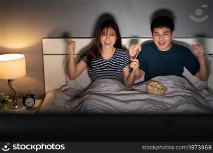 cheerful young couple watching sport TV with arm raised on a bed at night