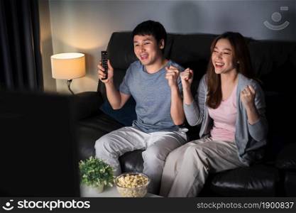 cheerful young couple watching sport TV with arm raised on sofa at night