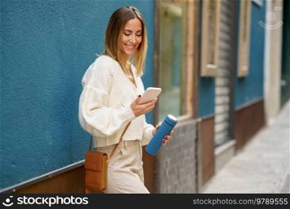 Cheerful young female in smart casual wear standing near blue wall while browsing mobile phone and messaging. Smiling woman with reusable bottle using smartphone on street