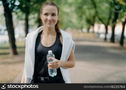 Cheerful young female runner holds water bottle poses outdoor in active wear takes break after workout walks in park gets refreshment after jogging feels thirsty keeps fit with regular sport