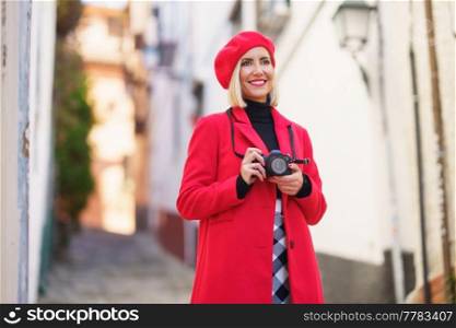 Cheerful young female traveler in fashionable red coat and beret, smiling and looking away, while standing on narrow street between residential buildings and taking photos on camera. Happy female tourist taking photos of town streets