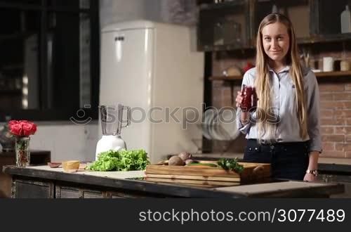 Cheerful young female with long blonde hair holding mason jar of fresh beet smoothie with striped straw and mint leaves for decoration in grunge styled kitchen and smiling. Organic food and healthy eating lifestyle.