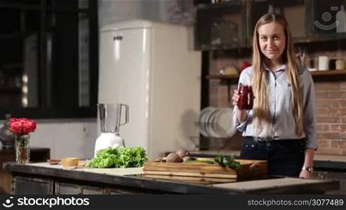Cheerful young female with long blonde hair holding mason jar of fresh beet smoothie with striped straw and mint leaves for decoration in grunge styled kitchen and smiling. Organic food and healthy eating lifestyle.