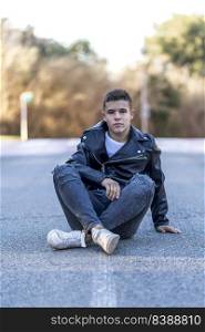 Cheerful young happy boy teenager sitting on asphalt road in front of the camera in posed portrait