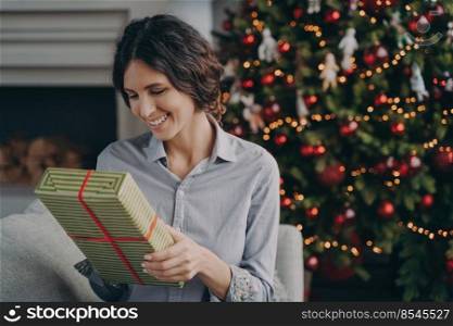Cheerful young Italian woman sitting on cozy sofa next festive decorated Xmas tree holding in hands Christmas present while looking at gift with broad sincere smile rejoicing coming winter holidays. Happy young Italian woman sits on cozy sofa next festive decorated Xmas tree with Christmas present