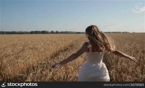 Cheerful young long blond hair woman in summer dress running through wheat field in summer during vacation. Joyful girl having fun, enjoying nature, freedom and happiness in countryside with beautiful landscape on background. Slo mo. Stabilized shot.