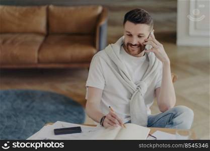 Cheerful young man freelancer or male entrepreneur talking with client on mobile phone and making some notes in notebook while working remotely from home, sitting in living room with leather sofa. Portrait of smiling man in white t-shirt covered with sweater, sitting on chair and talking on phone