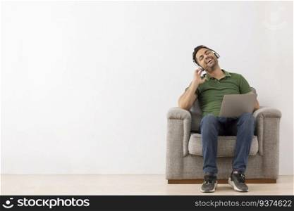 Cheerful young man listening music on headphones with eyes closed while sitting on sofa
