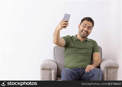 Cheerful young man taking selfie by using Smartphone while sitting on sofa