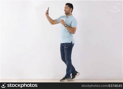 Cheerful young man taking selfie on Smartphone while standing with Coffee against white background