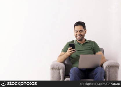 Cheerful young man using Smartphone while sitting on sofa with laptop on his lap