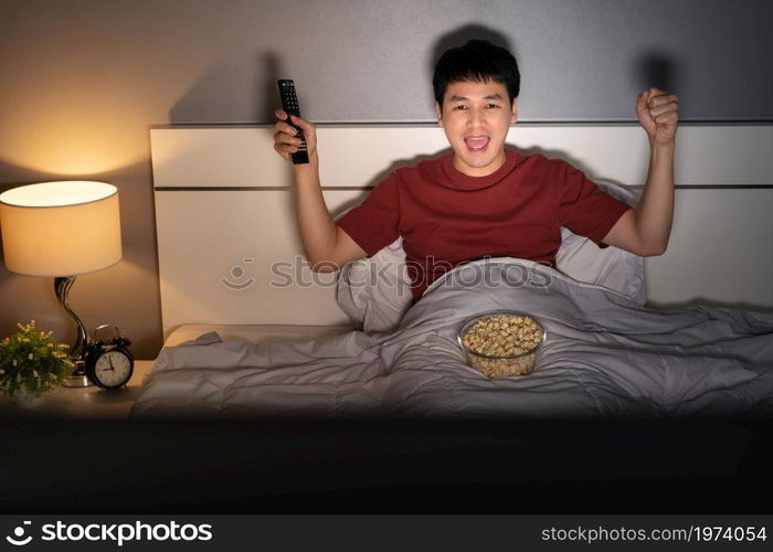 cheerful young man watching sport TV with arm raised on a bed at night