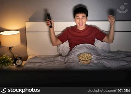 cheerful young man watching sport TV with arm raised on a bed at night