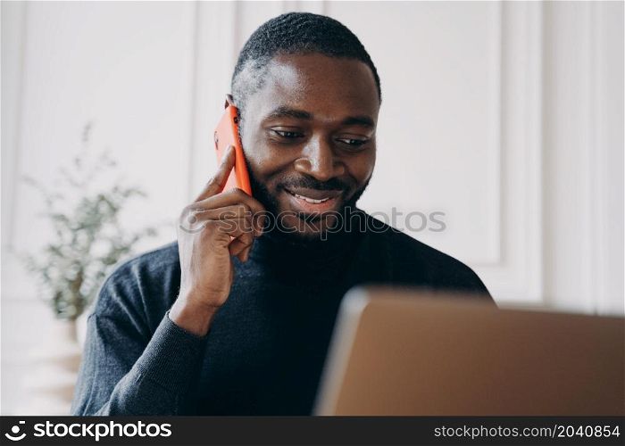 Cheerful young man with African lineage Ecommerce business owner calling to supplier for confirmation of orders while working remotely online from home office comparing and checking products on laptop. Cheerful young man with African lineage commerce businessowner calling to supplier for confirmation
