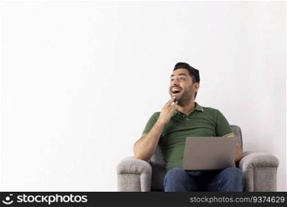 Cheerful young man with hand on chin looking away while using laptop on sofa 