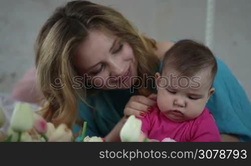 Cheerful young mother cuddling her cute baby girl and relaxing in bedroom. Adorable infant child playing with basket of tulip flowers while happy spending leisure at home. Slow motion. Stedicam stabilized shot.