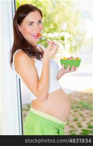 Cheerful young mother eating fruits, beautiful pregnant woman on the kitchen having healthy tasty lunch, new life concept