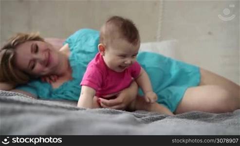 Cheerful young mother lying in bed and playing with her cute baby girl while enjoying sunny morning in a modern home bedroom. Happy mom embracing and kissing her smiling infant child while family relaxing at home. Slow motion. Stabilized shot.