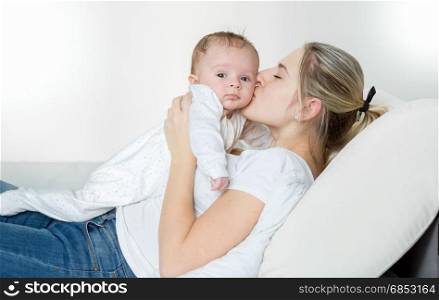Cheerful young mother lying on bed and kissing her 3 months old baby