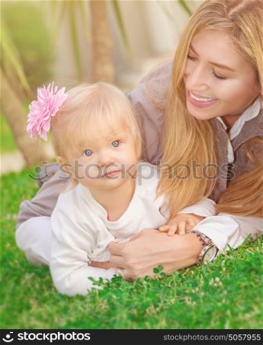 Cheerful young mother playing with her adorable little daughter in the park, lying down on fresh green grass field, happy family life