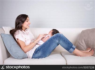 Cheerful young mother sitting on the couch at home and playing with cute little baby, happy motherhood, love and happiness concept