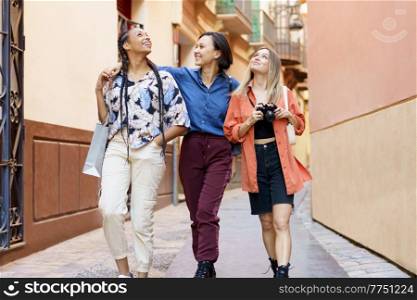 Cheerful young multiethnic female travelers in stylish clothes smiling and looking up while sightseeing old town during walk on street with photo camera. Happy multiracial female best friends walking in city during holidays