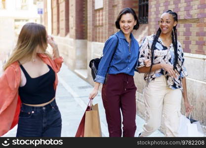 Cheerful young multiracial female millennials smiling happily while walking in city and chatting after successful shopping. Positive young diverse ladies smiling and strolling in city after purchases