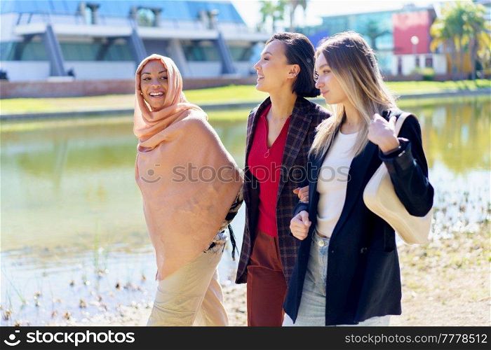 Cheerful young multiracial women friends in stylish clothes smiling and chatting while walking together on embankment near river on sunny day in city. Content young multiethnic girls talking while strolling on embankment in city