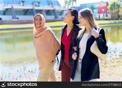 Cheerful young multiracial women friends in stylish clothes smiling and chatting while walking together on embankment near river on sunny day in city. Content young multiethnic girls talking while strolling on embankment in city