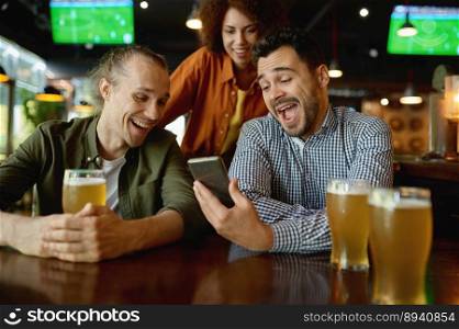 Cheerful young people rest in sport bar. Laughing friends sitting at table and watching funny video on smartphone while drinking draft beer. Cheerful young people rest in sport bar at table and watching video on phone