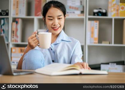 Cheerful young pretty woman sitting and use laptop computer and textbook to work or learning online, holding coffee mug in hand and smile with happy