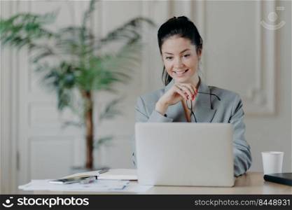 Cheerful young professional woman searches update notification for laptop computer, takes off glasses, dressed in formal clothes office interior. Freelancer works remotely searches ideas for blog site