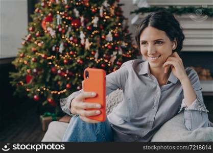 Cheerful young spanish woman enjoying christmas time, making selfie while sitting on comfy sofa next to festive bright colored Xmas tree. Pleasant blogger showing New Year time preparation at home. Cheerful young spanish woman enjoying christmas time at home, making selfie on smartphone