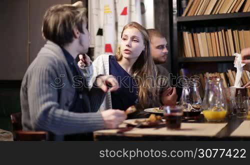 Cheerful young teenage friends enjoying meal together in grunge cafe. Beautiful teenage girl sharing salad with her boyfriend while spending great time together with groupmates at the restaurant.