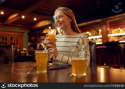 Cheerful young woman drinks coctail in bar. Group of people relax in pub, night lifestyle, friends celebrate event in restaurant. Cheerful young woman drinks coctail in bar