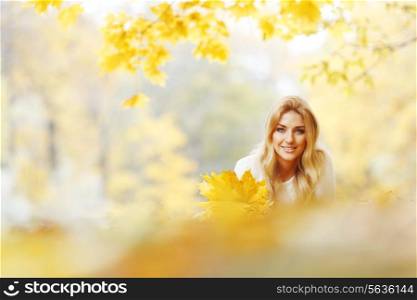Cheerful young woman laying down on the ground covered with dry autumnal foliage in beautiful park