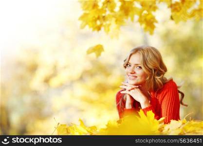 Cheerful young woman laying down on the ground covered with dry autumnal foliage in beautiful park