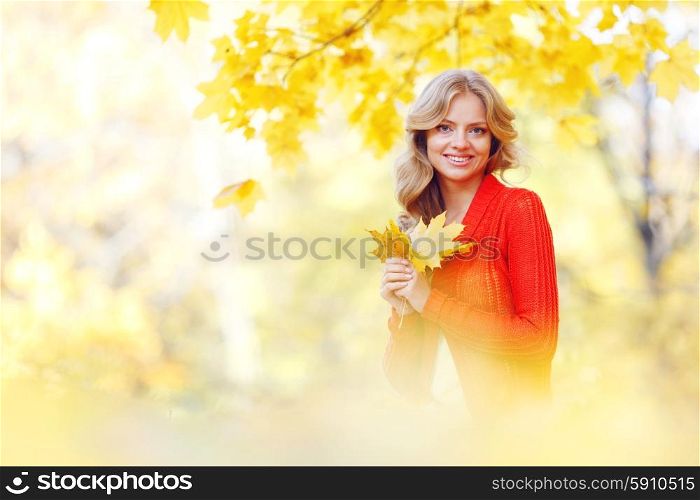 Cheerful young woman sitting on the ground covered with dry autumnal foliage in beautiful park